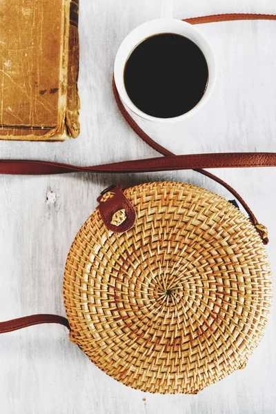 Bamboo bag, book and cup of coffee on white wooden background. Summer fashion, holiday