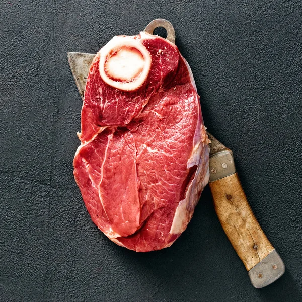 Piece of fresh beef meat on bone on dark stone background with butcher knife, top view