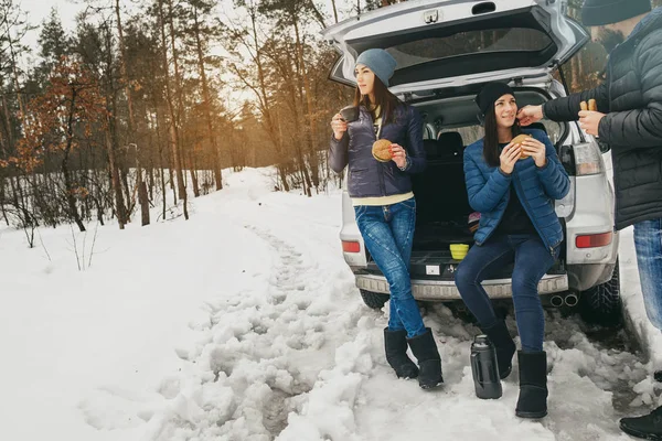 Group friends in winter wear on snowy day in winter forest drinking coffee in  car