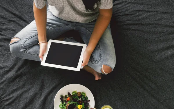 Woman holding digital tablet with empty blank screen and sitting on bed with salad