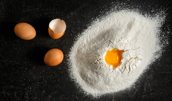 Heap of flour with eggs and yolk on the black background top view