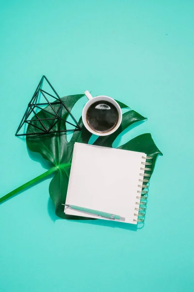 Summer background concept. Blank journal with monster leaves, pencil and cup of coffee on turquoise background. Top view flat lay