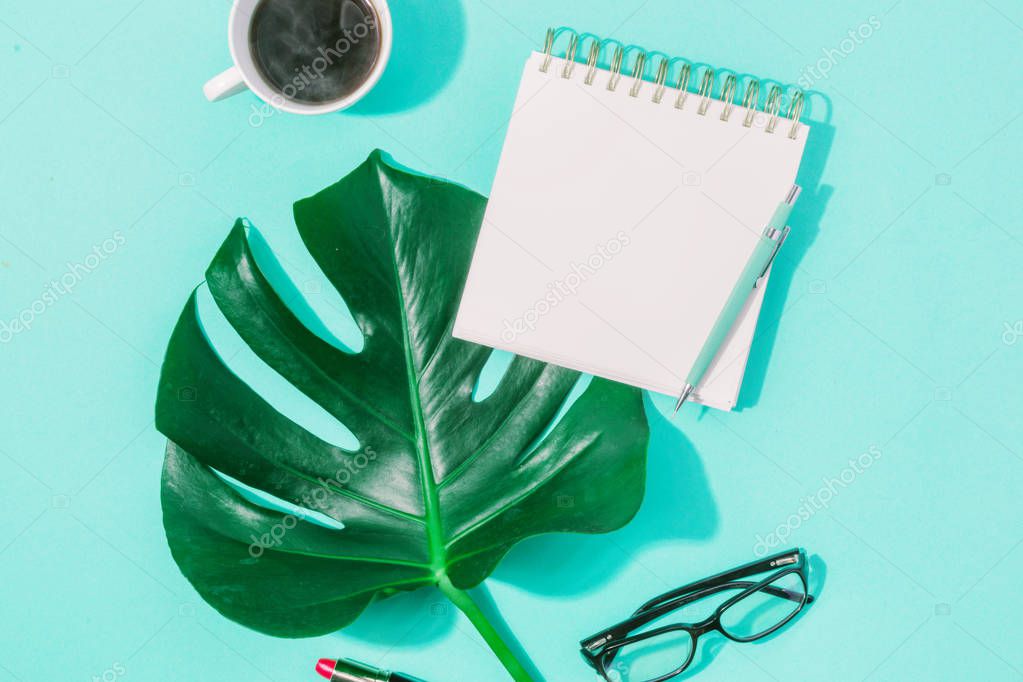 Summer background concept. Blank journal with monster leaves, pencil and cup of coffee on turquoise background. Top view flat lay