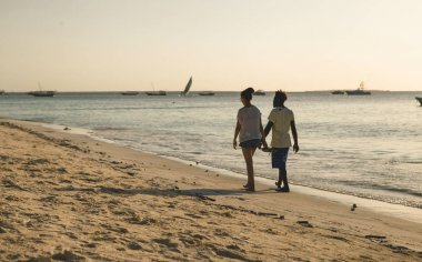 Birbirine aşık çiftler gün batımında okyanus kıyısında el ele yürüyorlar.