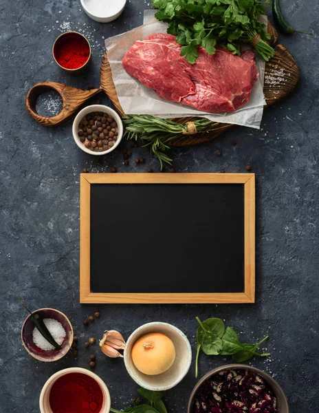 Cooking table with blank chalk board and beef meat, vegetables, spices, herbs top view. Food menu