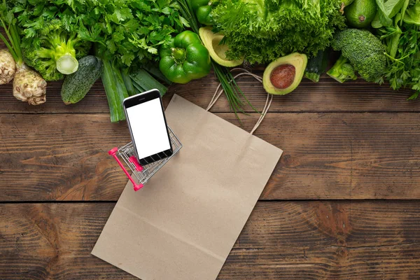 Order food online Phone in shopping cart with mock up on wooden background with clean green vegetables and paper bag top view