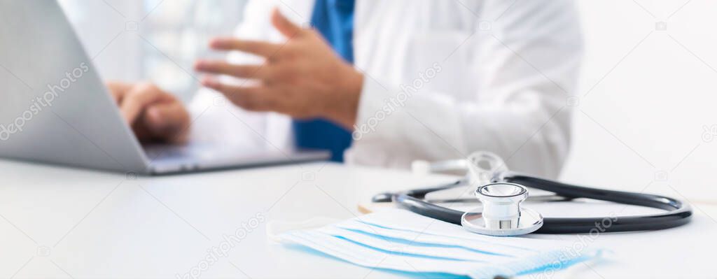 Online medicine concept. Stethoscope and medical mask on the doctors workplace in the background, the doctor conducts an online patient consultation using laptop