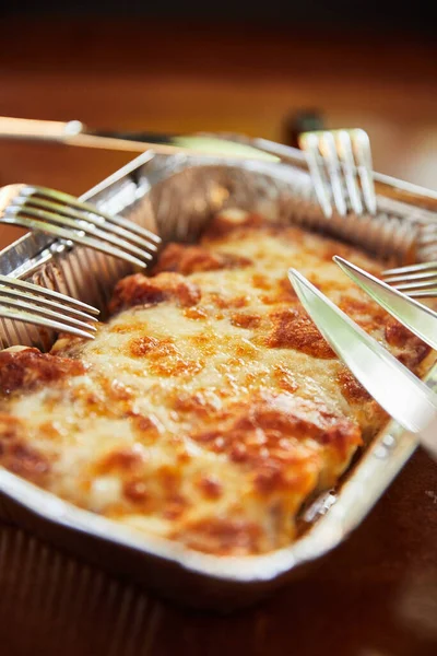 baked cheese dish in disposable rectangular tin paper aluminum foil bowl with forks and knives, close view