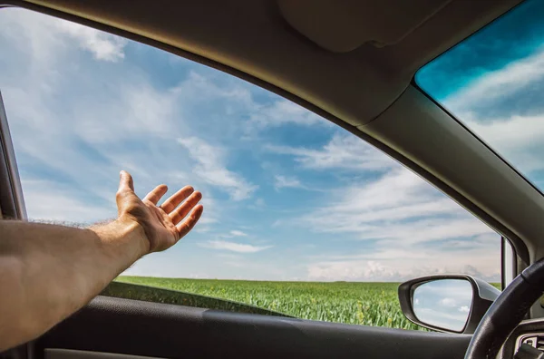 a man stuck his hand out of a car window while driving in sunny weather among green fields