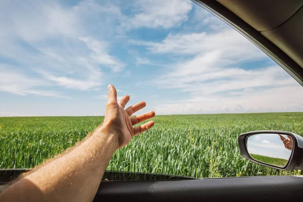 a man stuck his hand out of a car window while driving in sunny weather among green fields