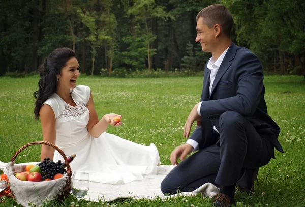 A couple of young people celebrate a wedding in a park on a picnic with wine and fruits, laugh and admire each other.
