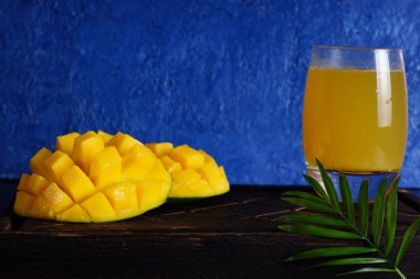 Mango ve suyu bir bardak parlak mavi arka plan ile ahşap bir gemide dilimlenmiş.
