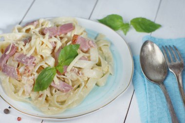 Yakınındaki bir ahşap beyaz masa üzerinde duran tabak, çatal ile bir tabakta Fesleğenli makarna carbonara