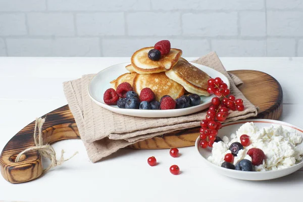 On the table is a wooden board with pancakes with fresh berries of currant, blueberry and raspberry, and next to a bowl of cottage cheese — Stock Photo, Image