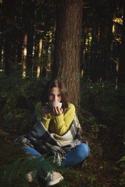 Ekose sarılmış genç bir esmer kız bir ağacın yanında bir ormanda oturur ve büyük bir kupa çay içer