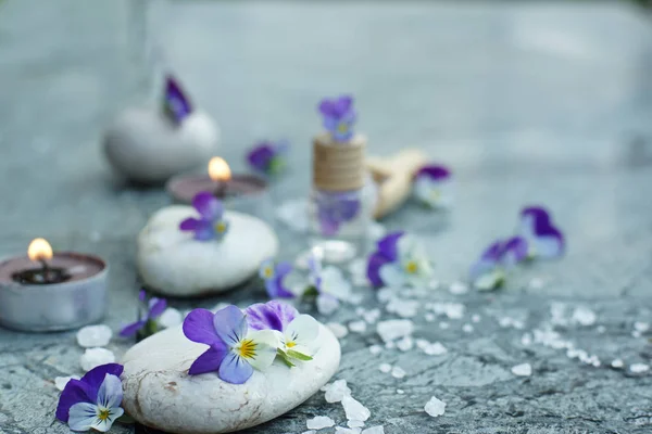 Zacht stilleven met violette viooltjes en rekwisieten voor spabehandelingen, wit badzout, brandende kaarsen en stenen voor een warme massage — Stockfoto