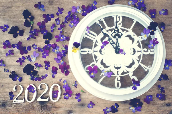 Figuras 2020 sobre un fondo de madera junto a flores de color púrpura y un reloj blanco que muestra cinco minutos hasta la medianoche — Foto de Stock