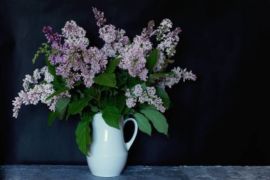 Büyük bir demet mor leylak, siyah arka planda beyaz bir sürahi içinde.