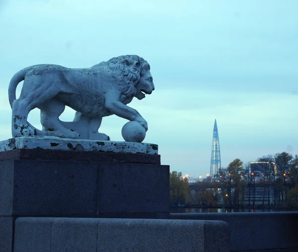 Uma escultura de um leão em um pedestal de granito um leão que prende uma bola com sua pata nos edifícios da distância e nas estruturas da cidade de São Petersburgo na Rússia pode ser vista Imagens Royalty-Free