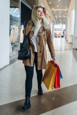 mutlu kız gülümseme ve büyük şehir alışveriş merkezi beyaz çanta alışveriş çok renkli yürüme. Genç Sarışın mutlu bir gülücük ver paketleri alışveriş ile büyük mağaza. Alışveriş, satış, tüketim ve yaşam tarzı kavramı