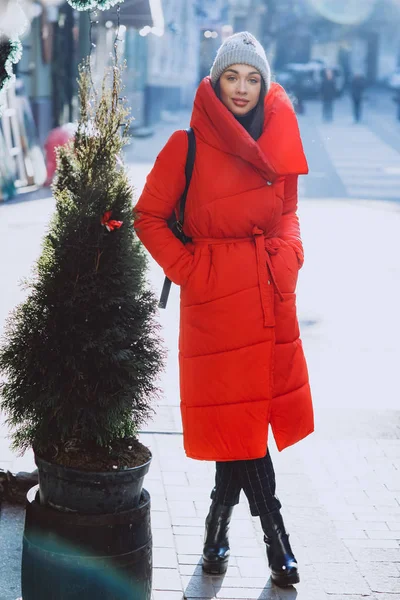 在新年街道上 穿着红色羽绒服和灰色针织帽子的漂亮年轻女子的生活方式时尚肖像停留在圣诞树附近 圣诞节 新年概念 — 图库照片