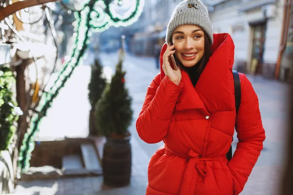 愉快的女人穿着红色夹克和针织帽子在街上用手机说话 圣诞节 新年概念 — 图库照片