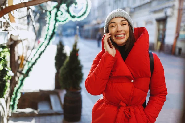 一个相当快乐的女人 一个雪微笑着白色的牙齿 穿着红色外套和针织帽子在智能手机上交谈 新年和圣诞节假期 冬季概念 — 图库照片