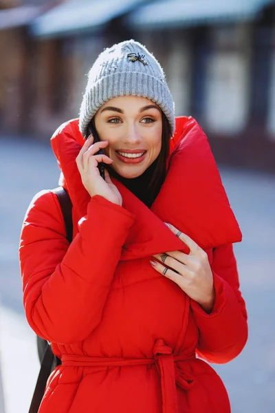 快乐的时尚女性使用智能手机 在冬季的街道上穿着红色外套 如羽绒服和灰色针织帽子说话 寒假与交流 — 图库照片