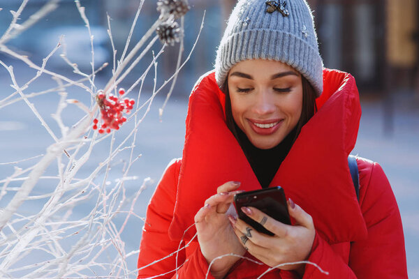 Smilling Girl in red winter jacket types something in her phone standing on the winter street near traditional christmas tree.  Christmas, new year and winter holiday concept - Image