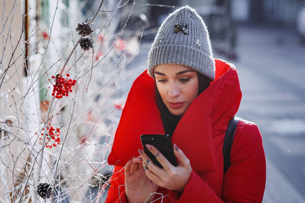 close up portrait of happy woman in red winter down jacket and knitted grey hat types text of sms or message  in her mobile smartphone standing on the winter street near traditional christmas tree.  Christmas, new year and winter holiday concept - Im
