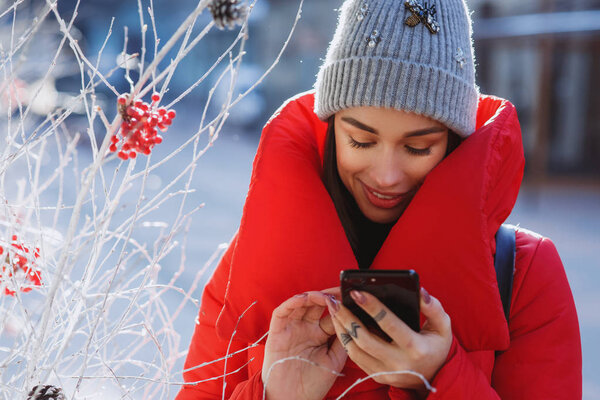 close up portrait of happy woman in red winter down jacket and knitted grey hat types text of sms or message  in her mobile smartphone standing on the winter street near traditional christmas tree.  Christmas, new year and winter holiday concept - Im