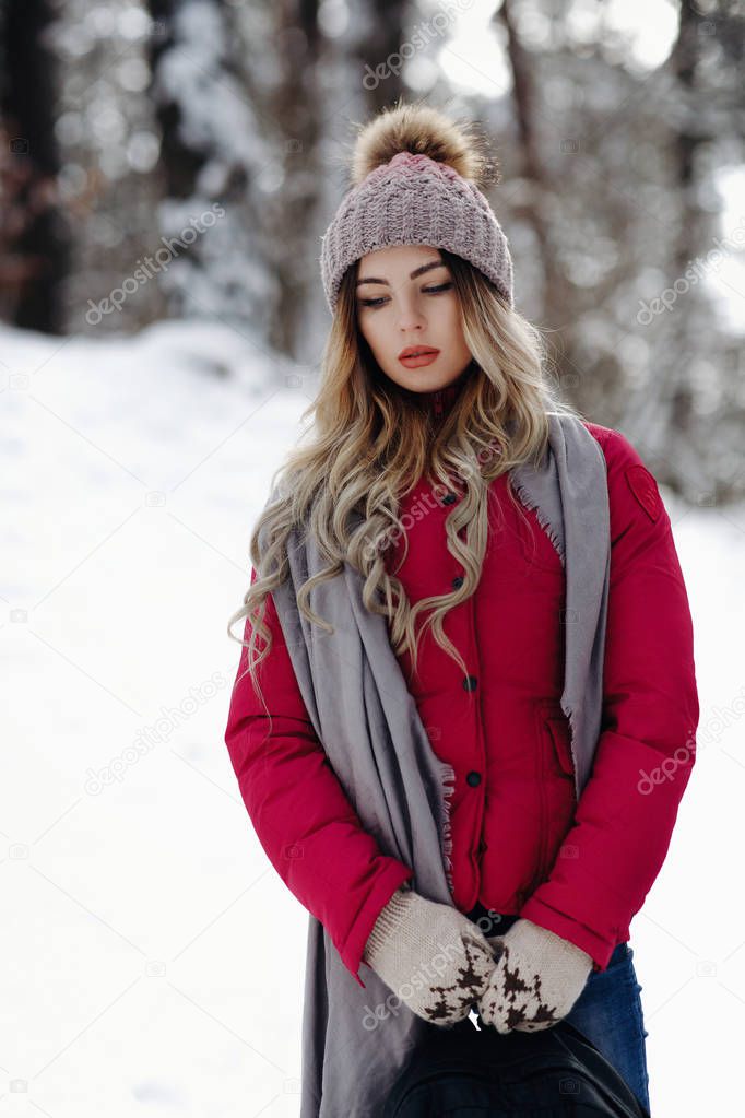 beautiful stylish girl enjoying wonderful winter snowfall in forest park standing outside and hold bag in gloves wearing knitted hat whil ball, grey scarf and red jacket snowy weather flying snowflakes day beauty fashion casual clothes woman good moo