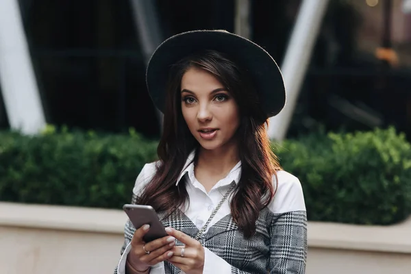 Portret van stijlvolle vrouw met moderne zwarte hoed kijken opzij met behulp van mobiele telefoon — Stockfoto
