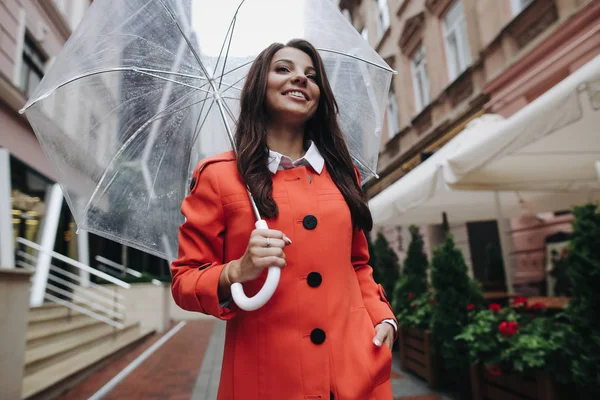 Woman with umbrella wearing red coat in a good mood. Portrait of beautiful brunette girl with umbrella rainy day standing near the cafe. — Stock Photo, Image