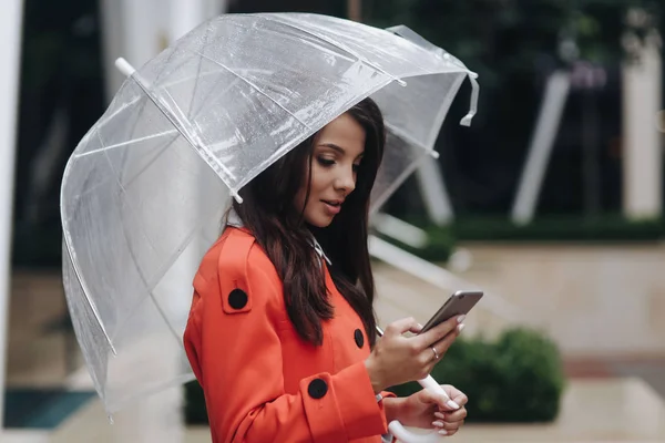 Mooie brunette vrouw met paraplu regenachtige dag in rode vacht typen Tekstbericht op mobiele telefoon en staande in de buurt van Boutique. — Stockfoto