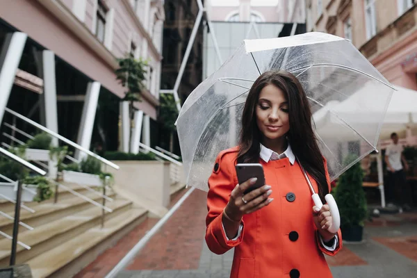 Beautiful woman with umbrella rainy day in red coat dialing number on phone making video call using smart phone and standing near old building.