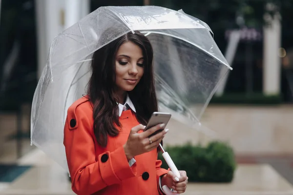 Mooie brunette vrouw met paraplu regenachtige dag in rode vacht typen Tekstbericht op mobiele telefoon en staande in de buurt van Office Center. — Stockfoto