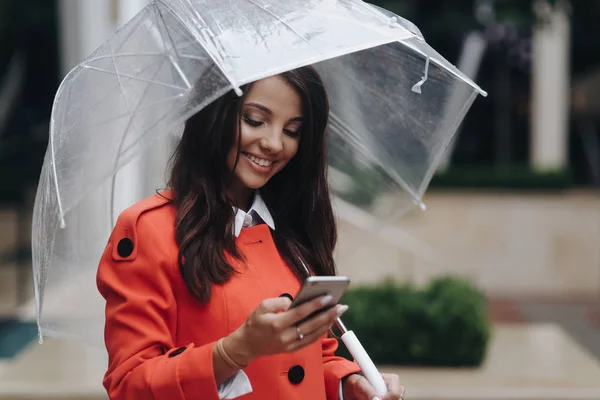 Красивая улыбающаяся женщина с зонтиком дождливый день в красном пальто, глядя внимательно на экран мобильного телефона и стоя рядом с офисным центром . — стоковое фото