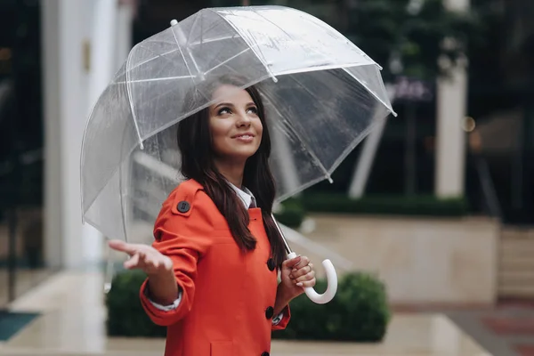 Portret van een langharige stijlvolle jonge meisje te wachten op regen in rode vacht kijken naar de hemel op de achtergrond van de stad. — Stockfoto