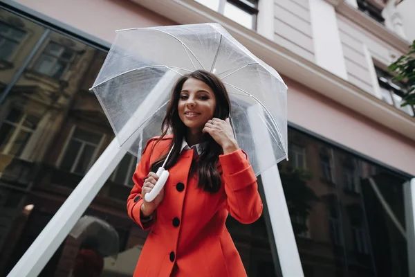 Portret van een langharige stijlvolle jong meisje met paraplu regenachtige dag in rode vacht kijken naar camera op de achtergrond van de stad — Stockfoto