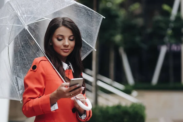 Vrouw schrijven SMS op straat met paraplu. Buiten portret van jonge mooie vrouw chating met vrienden, staande op straat. — Stockfoto