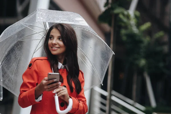 Vrouw houdt telefoon op straat met paraplu en kijkt opzij. Buiten portret van jonge vrouw chating met vrienden — Stockfoto