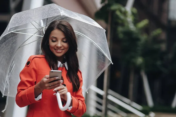Woman writing sms on the street with umbrella and smiling. Outdoor portrait of young beautiful woman chating with friends