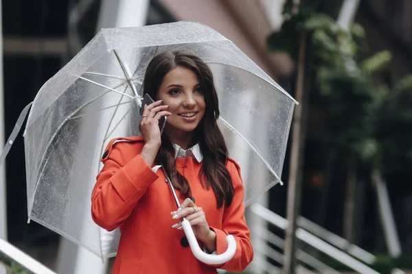 Mujer sosteniendo teléfono y paraguas en la calle, hablando por su teléfono. Retrato al aire libre de la joven mujer chating con amigos, de pie en la calle —  Fotos de Stock