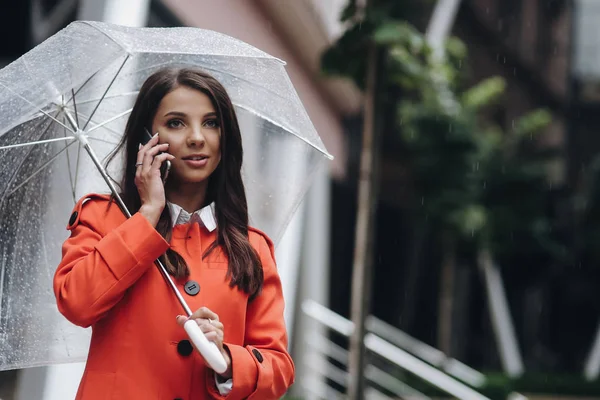 Brunette vrouw praten op telefoon houden paraplu en kijken opzij. Buiten portret van jonge vrouw praten met vriend op mobiele telefoon — Stockfoto