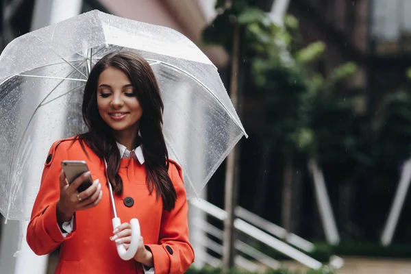 Lachende vrouw houdt paraplu lezing bericht op mobiele telefoon terwijl staan op de straat. — Stockfoto