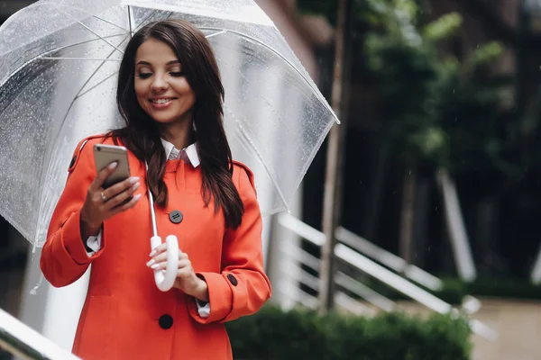 Modieuze jonge lachende vrouw bezig met haar mobiele telefoon terwijl staan in de buurt van de winkel, het houden van paraplu — Stockfoto