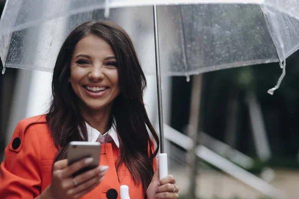 Портрет красивой улыбающейся женщины с прозрачным зонтиком, пишущей сообщение в городе . — стоковое фото
