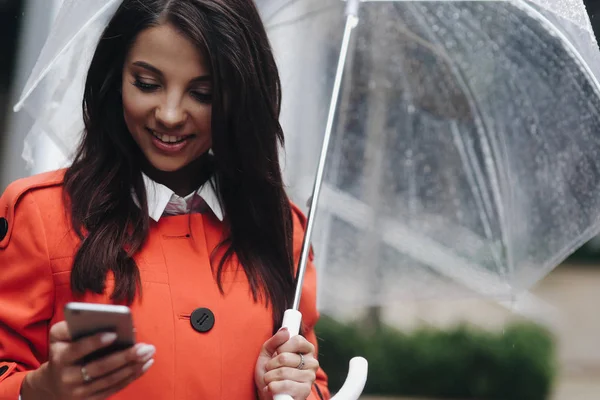 Portret van jonge vrouw met transparante paraplu schrijven SMS op telefoon in stad. — Stockfoto