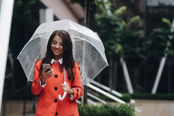 Mooie vrouw met transparante paraplu wandelen en lezen van SMS-berichten op op een slimme telefoon in de stad. — Stockfoto